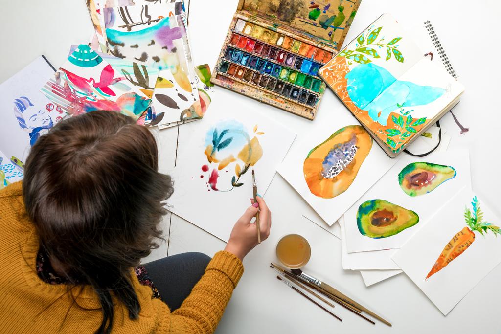Draufsicht der Frauenmalerei mit Aquarellfarben, während sie von Farbzeichnungen und Zeichenutensilien umgeben ist - Foto, Bild