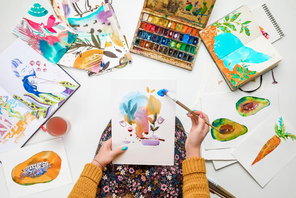 Widok góry kobietę, opierając się na kolana i malarstwo w nim z farby akwarele w otoczeniu kolorowych zdjęć - Zdjęcie, obraz