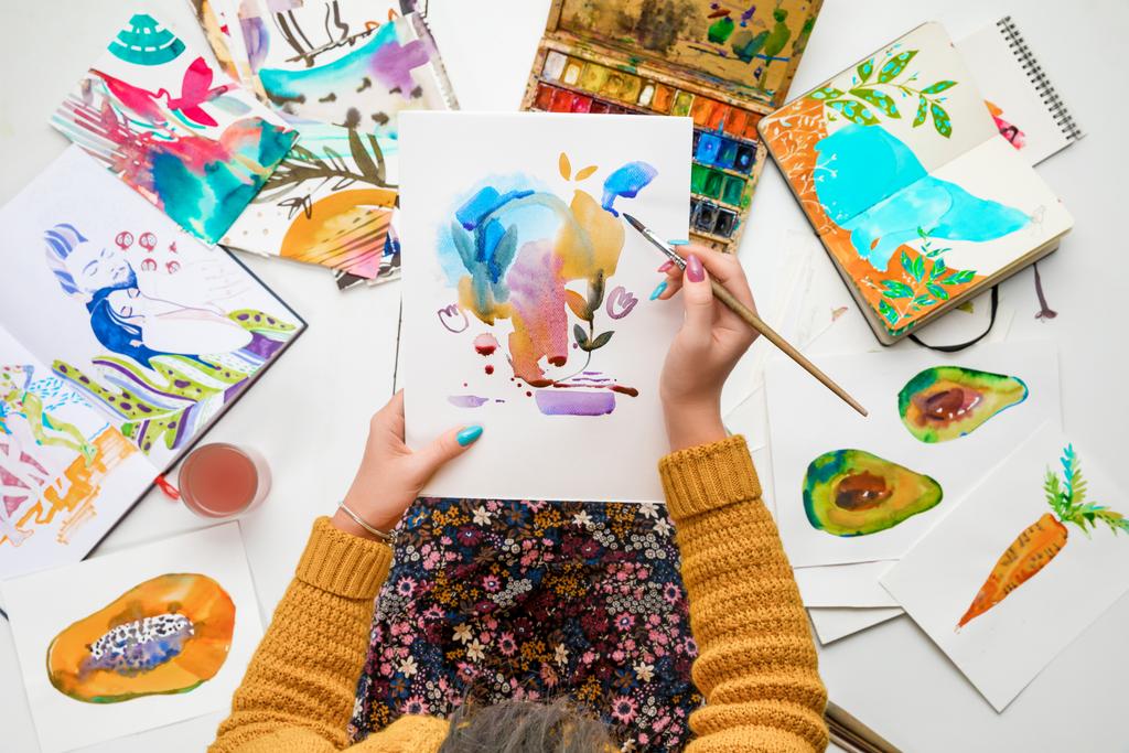 Draufsicht einer Frau, die eine Zeichnung auf Knien hält und sie mit Aquarellfarben bemalt, während sie von farbigen Bildern umgeben ist - Foto, Bild
