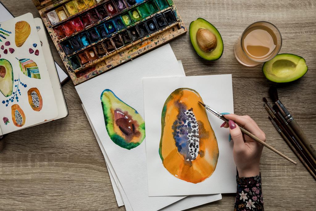 top view of female hands drawing avocado and papaya with watercolor paints, paintbrush on wooden table - Photo, Image