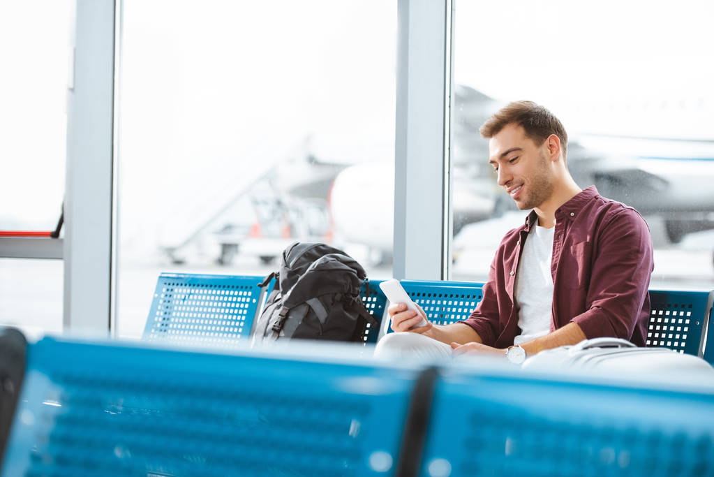 cheerful man holding smartphone while sitting in departure lounge - Photo, Image