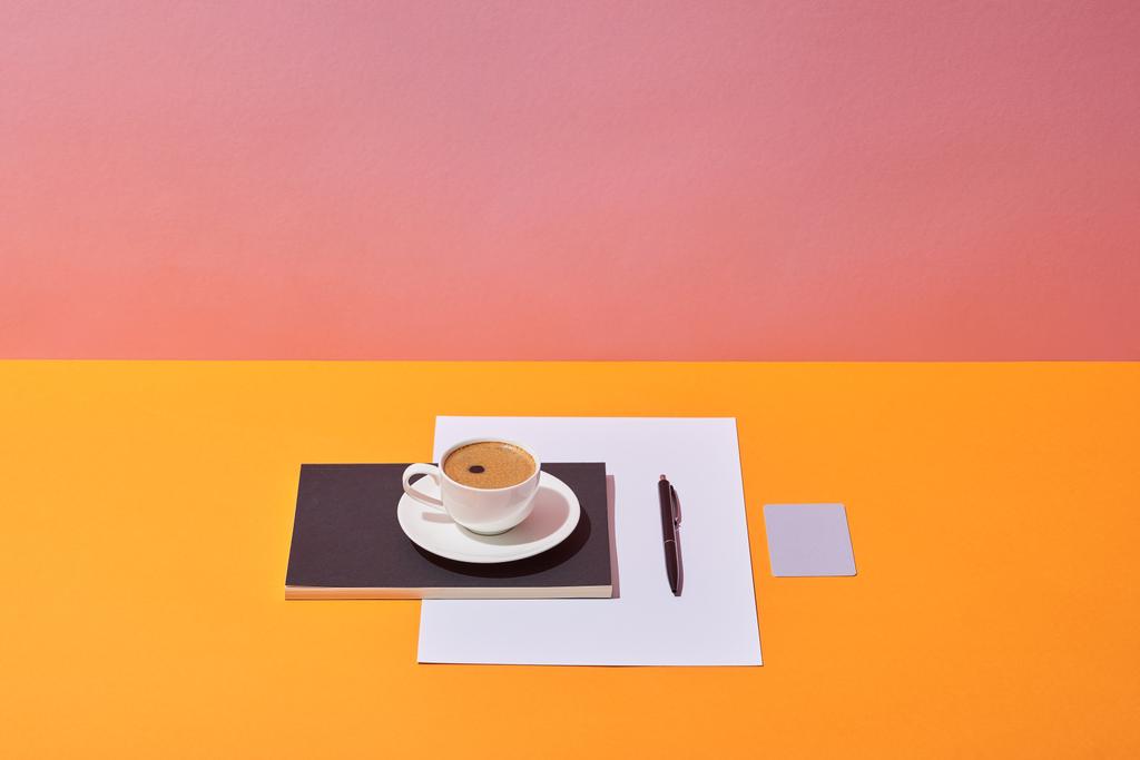 xícara de café, pires, caneta, folha de papel e caderno na mesa amarela e fundo rosa
 - Foto, Imagem