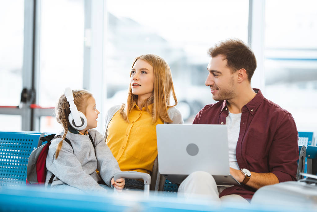 cute kid in headphones looking at mother while sitting near father in airport  - Photo, Image