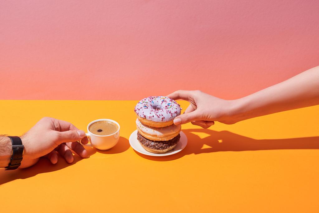 περικοπεί προβολής της γυναίκας και άνδρα, λαμβάνοντας νόστιμα ντόνατς στο πιατάκι και το φλιτζάνι του καφέ στην κίτρινη επιφάνεια και ροζ φόντο - Φωτογραφία, εικόνα