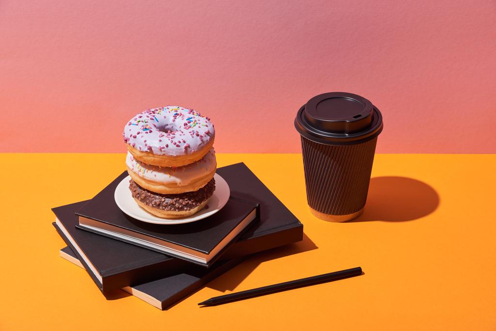 tasty donuts on saucer and paper coffee cup near notebooks and pencil on yellow desk and pink background - Photo, Image