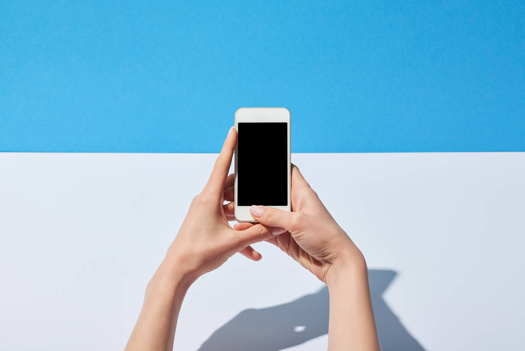 περικοπεί προβολή της γυναίκας χρησιμοποιώντας smartphone με κενή οθόνη στην άσπρη επιφάνεια και μπλε φόντο - Φωτογραφία, εικόνα