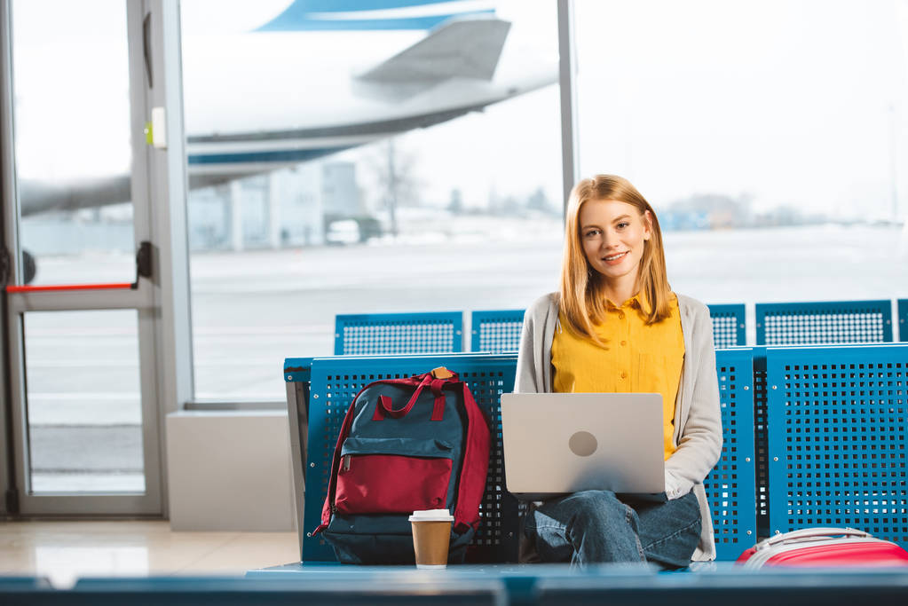 beautiful woman sitting with laptop near backpack and disposable cup in airport  - Photo, Image