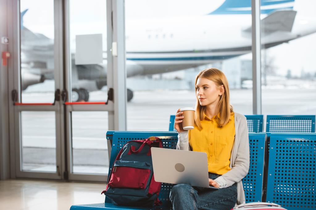 gyönyörű nő ül a laptop, és a gazdaság eldobható pohár a kezében, repülőtér  - Fotó, kép
