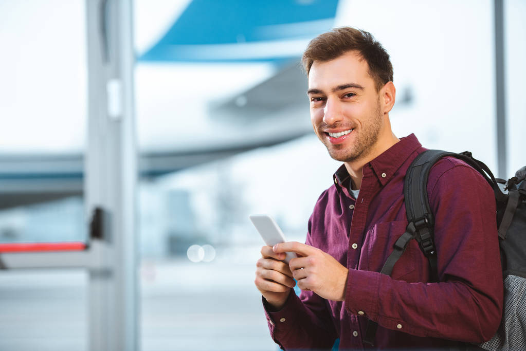 vidám ember gazdaság smartphone, és mosolyogva repülőtér  - Fotó, kép