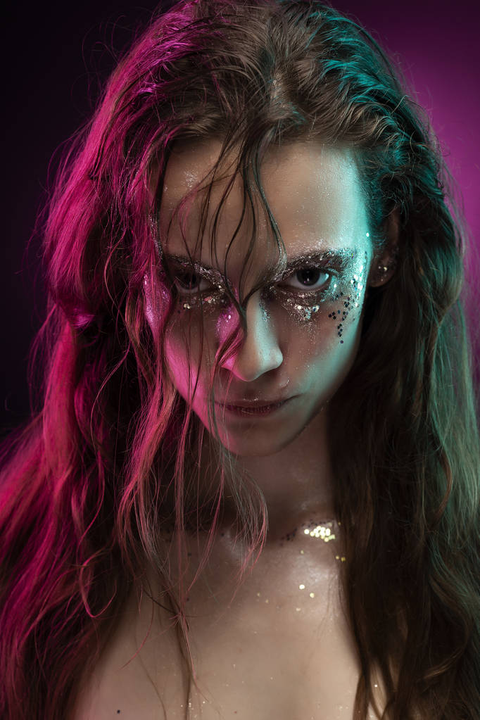 Mooi meisje met creatieve make-up gemaakt van glitter met tranen over haar gezicht verlicht met roze en blauw licht. Roze achtergrond. Conceptuele, reclame en commerciële design. Kopiëren van ruimte. - Foto, afbeelding