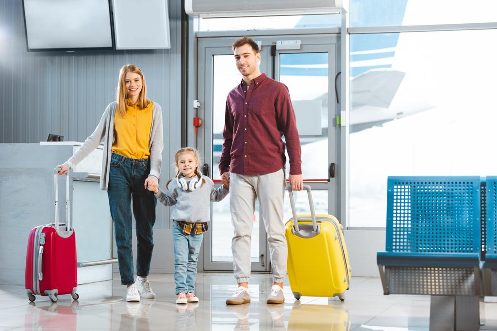 happy mom and dad holding hands with daughter and standing with suitcases near gate in airport - Photo, Image