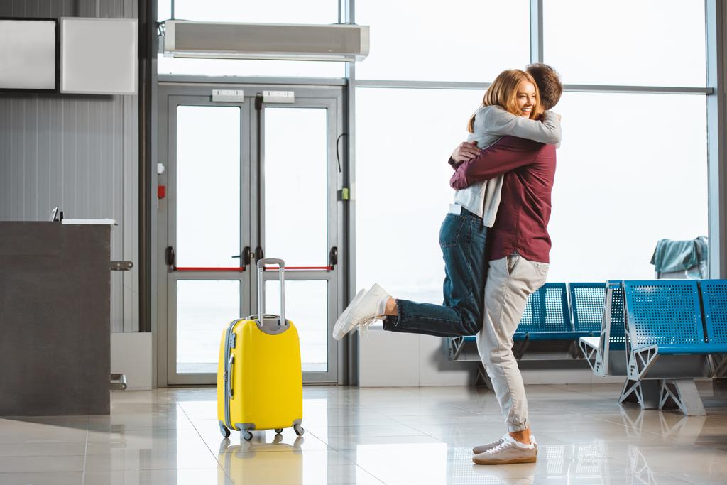 atractiva mujer abrazando novio en sala de espera cerca de la maleta
 - Foto, Imagen