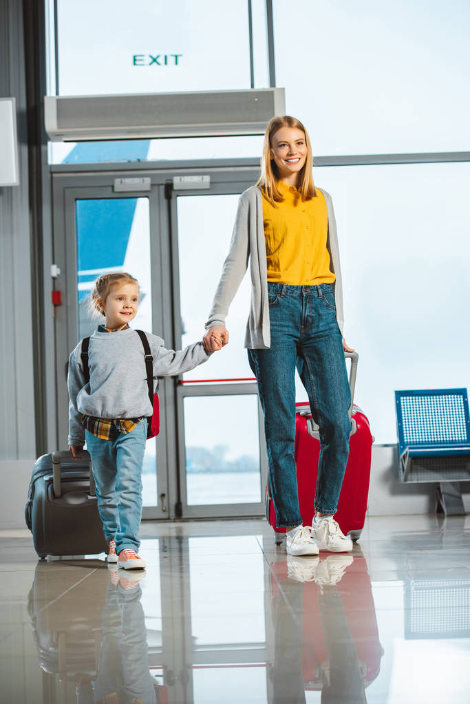 attractive mother holding hands with cute daughter while walking with suitcases in airport  - Photo, Image