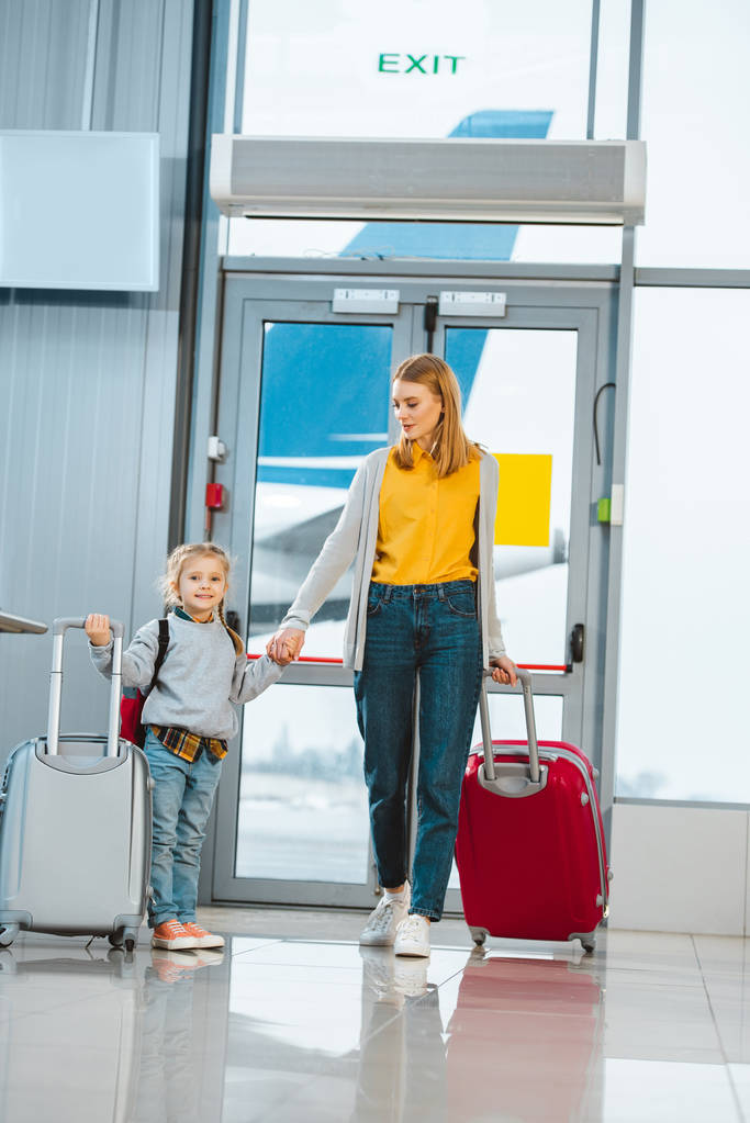 cheerful mother looking at daughter while holding hands in airport - Photo, Image