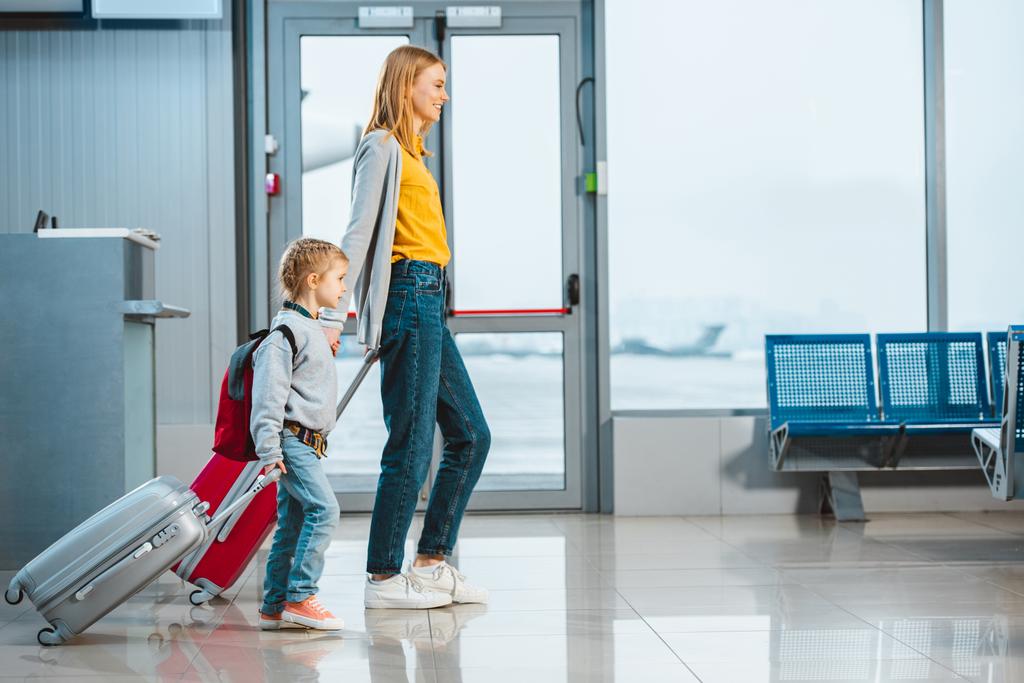 äiti ja tytär pitävät toisiaan kädestä ja kävelevät matkatavaroiden kanssa lentokentällä
  - Valokuva, kuva