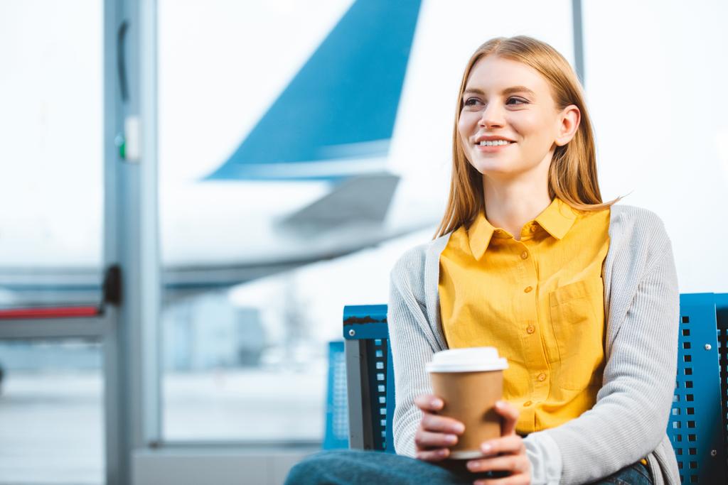 schöne Frau hält Pappbecher, während sie im Flughafen sitzt  - Foto, Bild