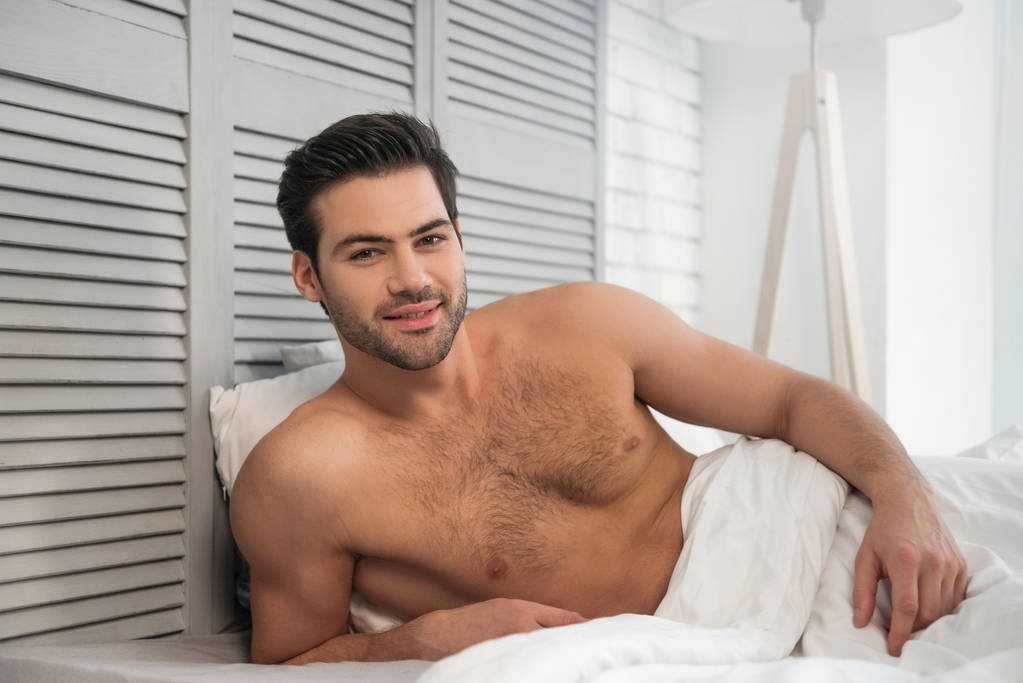 retrato de hombre desnudo sonriente mirando a la cámara acostado en la cama por la mañana
 - Foto, Imagen