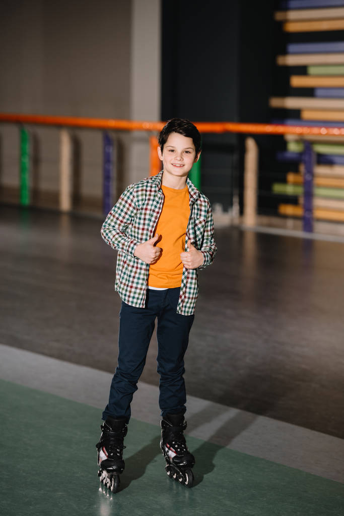Выборочный фокус милого улыбающегося мальчика в роликовых коньках, стоящего на просторном катке
 - Фото, изображение