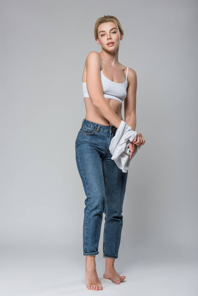Slim blonde meisje in jeans en witte beha uitkleden geïsoleerd op grijs - Foto, afbeelding