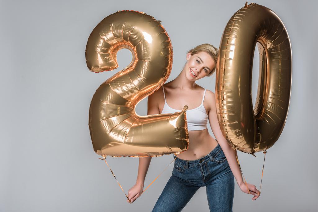 attractive smiling girl in jeans and white bra posing with 20 golden balloons, isolated on grey - Photo, Image
