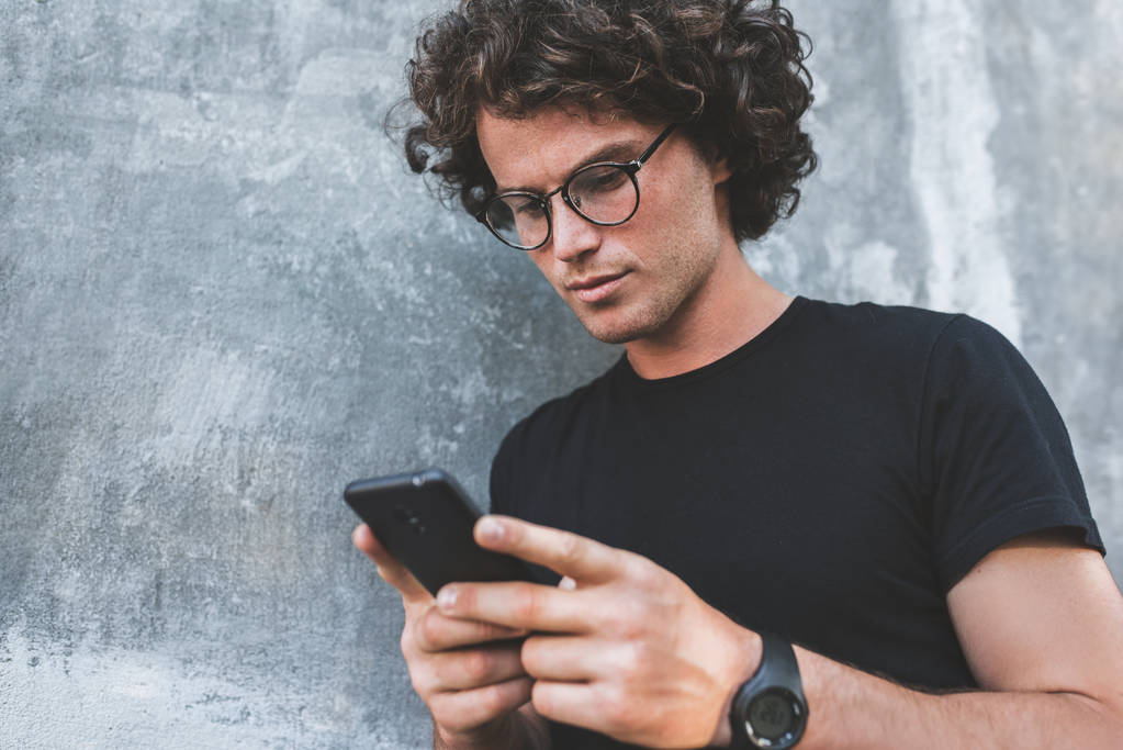 hombre guapo con pelo rizado sosteniendo el teléfono móvil mientras está de pie contra la pared gris
 - Foto, Imagen