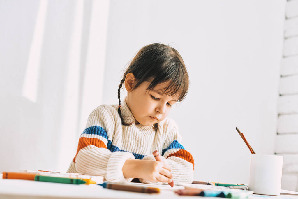 little girl painter painting with oil pencils, sitting at white desk at home - Photo, Image