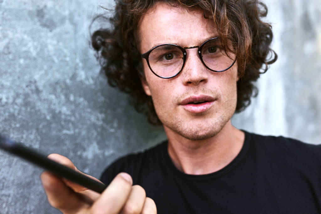 homem bonito com cabelo encaracolado olhando para a câmera, segurando telefone celular enquanto está contra a parede cinza
 - Foto, Imagem