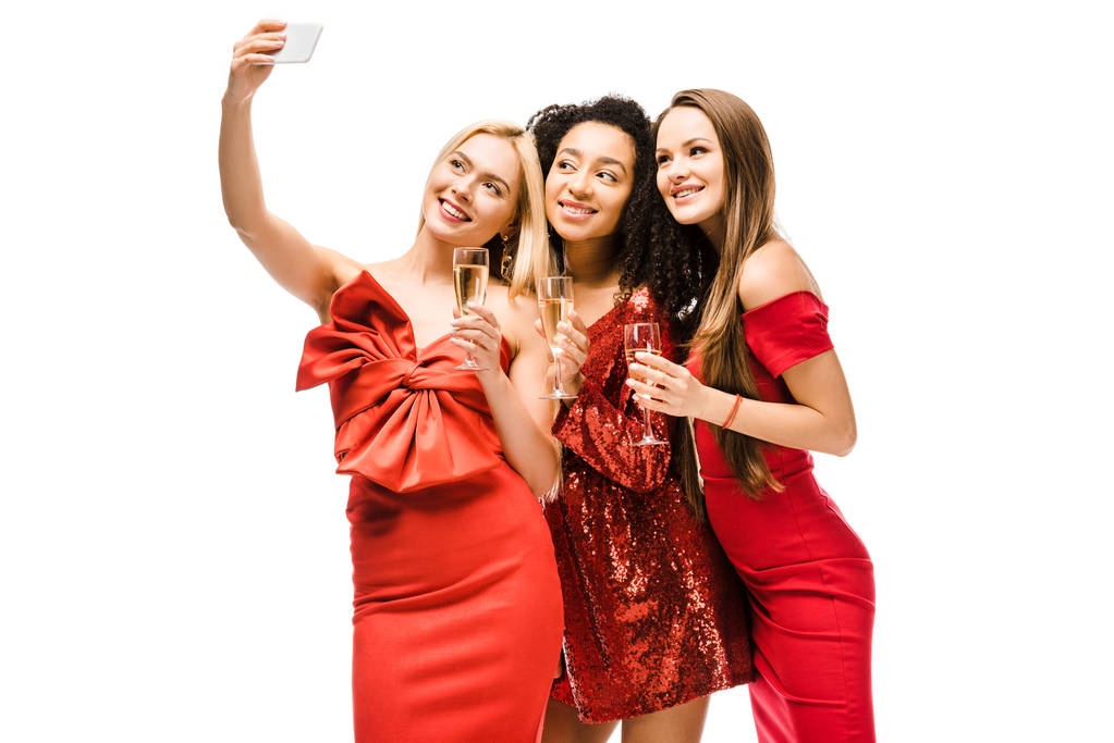 schöne multiethnische Mädchen in roten Kleidern mit Champagnergläsern machen Selfie auf dem Smartphone isoliert auf weiß - Foto, Bild