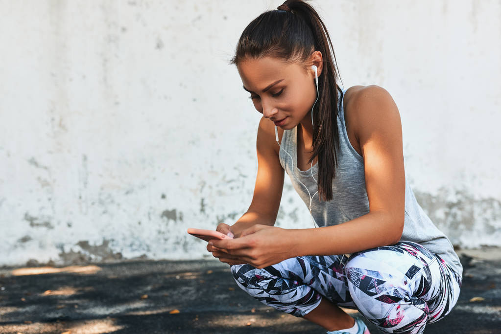 Fitness young pretty woman listening the music on earphones during a break outdoors against concrete wall. Athletic female relaxing after exercises sitting with smartphone, after workout outside. - Photo, Image