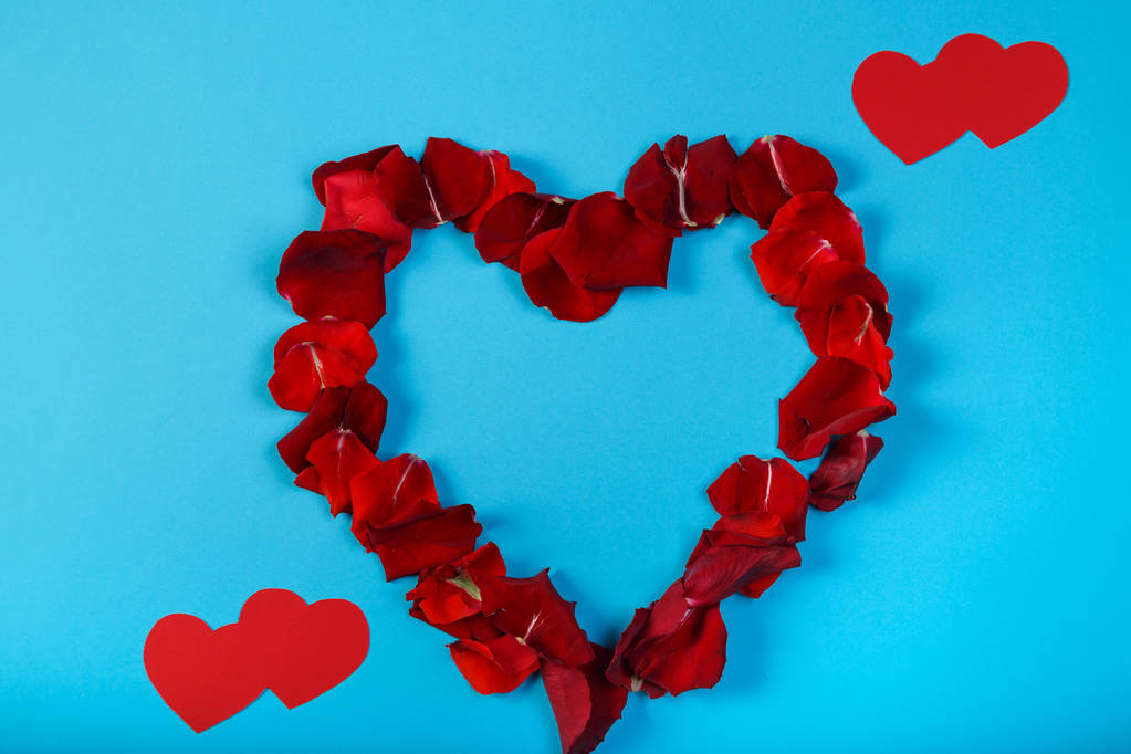 Een hart bekleed met roze bloemblaadjes op een blauwe achtergrond en rode papier harten. - Foto, afbeelding