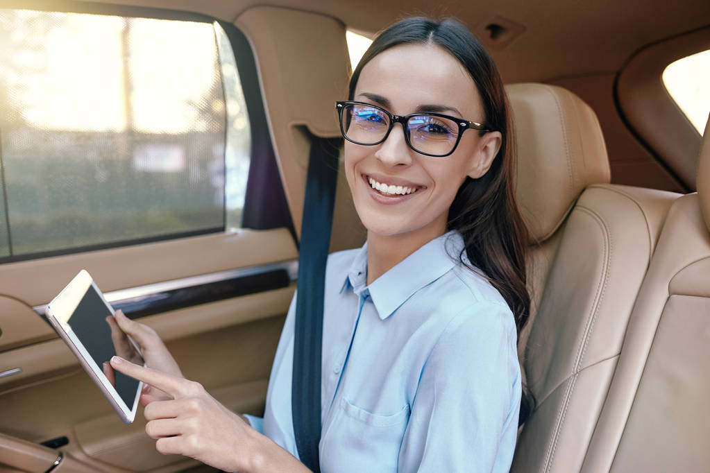 hymyilevä valkoihoinen liikenainen, joka käyttää digitaalista tablettia ja katsoo kameraa autossa
 - Valokuva, kuva