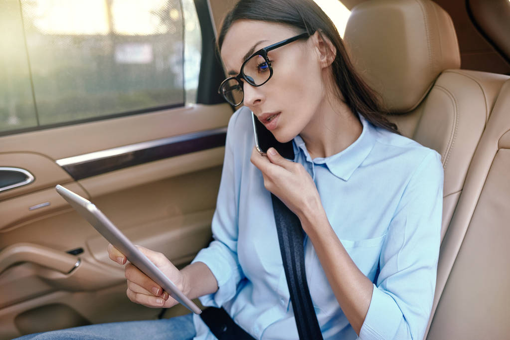portrait of businesswoman talking on smartphone and looking at digital tablet in the car - Fotoğraf, Görsel