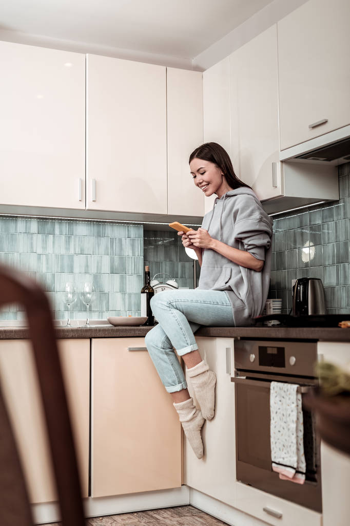 dunkelhaarige Frau in Jeans und übergroßem Kapuzenpulli sitzt in Küche - Foto, Bild