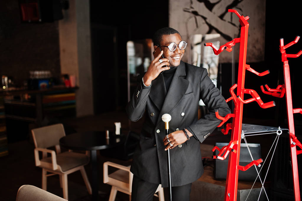 Élégant homme afro-américain dans une élégante veste noire et des lunettes, tenant rétro bâton de marche comme une fiole de canne ou une canne à bascule avec poignée en diamant doré et parlant sur téléphone portable
. - Photo, image