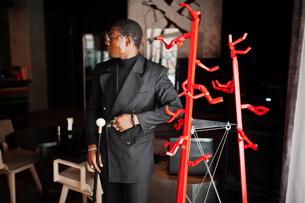 Élégant homme afro-américain dans une élégante veste noire et des lunettes, tenant bâton de marche rétro comme une fiole de canne ou une canne à bascule avec poignée en diamant doré. Riche homme afro à la mode
. - Photo, image