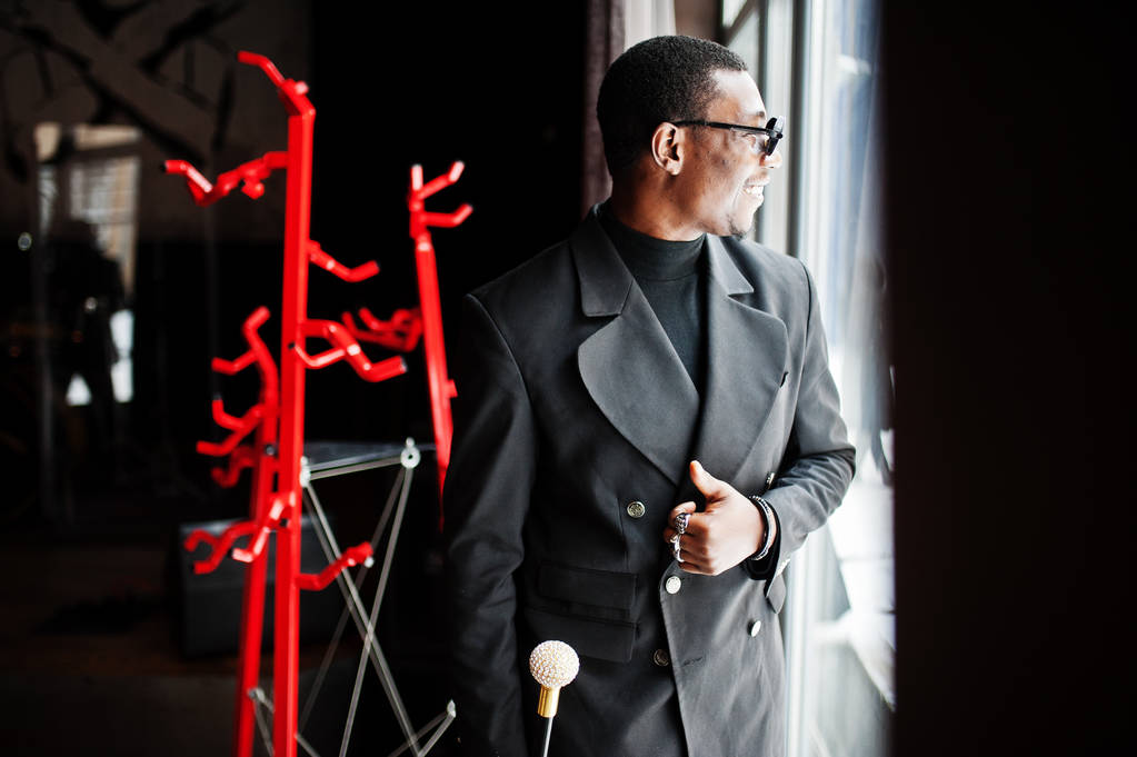 Стильный африканский американский джентльмен в элегантной черной куртке и солнцезащитных очках, держа в руках тростниковую фляжку или тростник с золотой бриллиантовой рукояткой. Богатый афро-человек
. - Фото, изображение