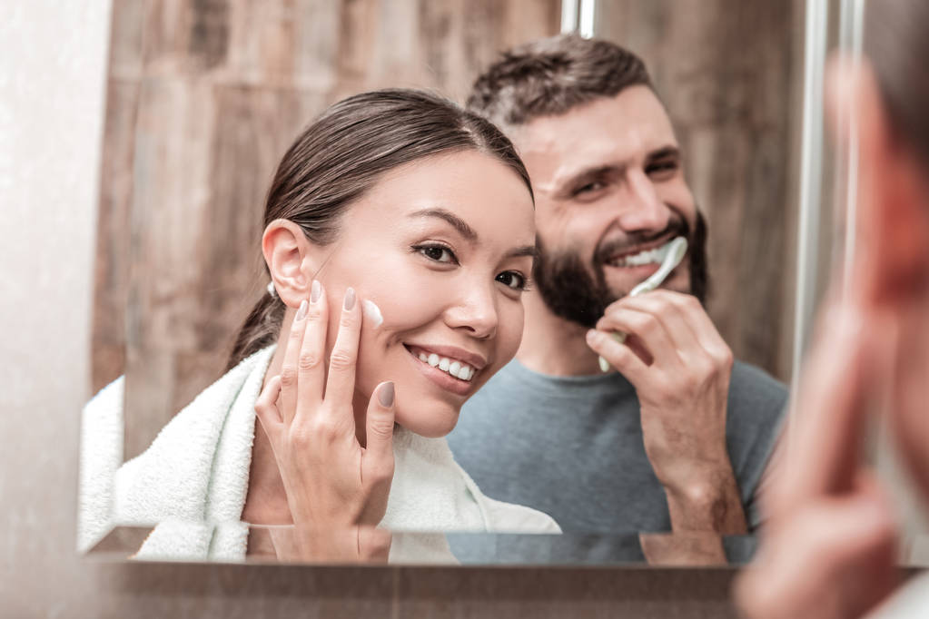 Kaunis nainen, joka käyttää kasvovoidetta, kun mies harjaa hampaita
 - Valokuva, kuva