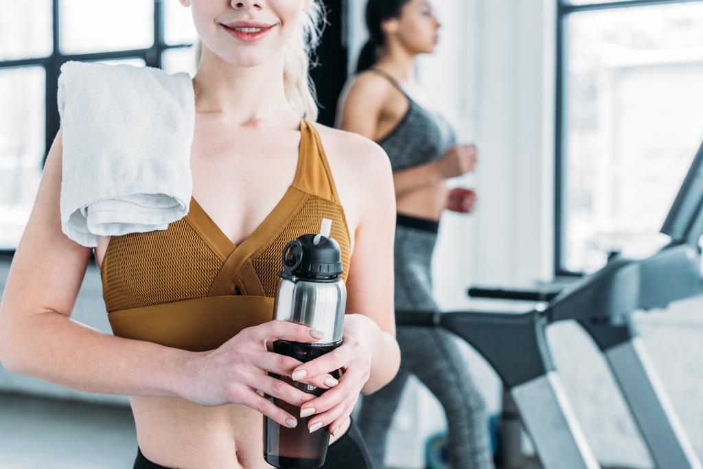cropped shot of smiling sporty girl with towel on shoulder holding sports bottle in gym - 写真・画像
