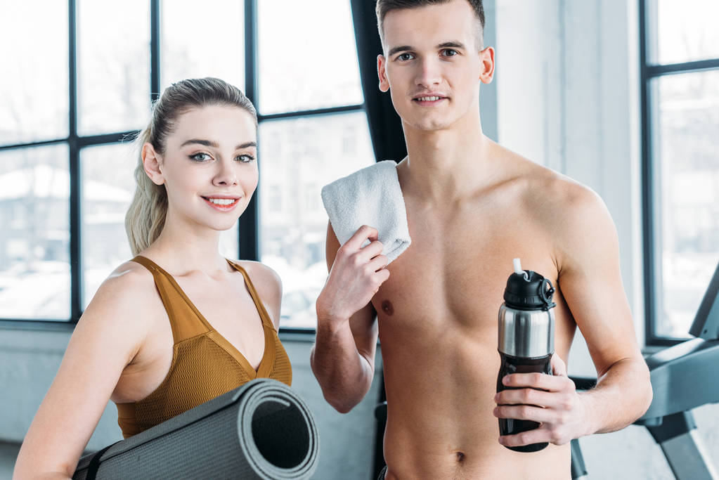 женщина с ковриком для йоги и мужчина без рубашки с полотенцем и спортивной бутылкой стоя вместе и улыбаясь на камеру в тренажерном зале
 - Фото, изображение