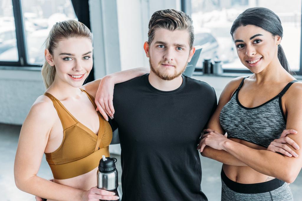 handsome male trainer with beautiful multiethnic sporty girls standing together and smiling at camera in gym - Photo, Image