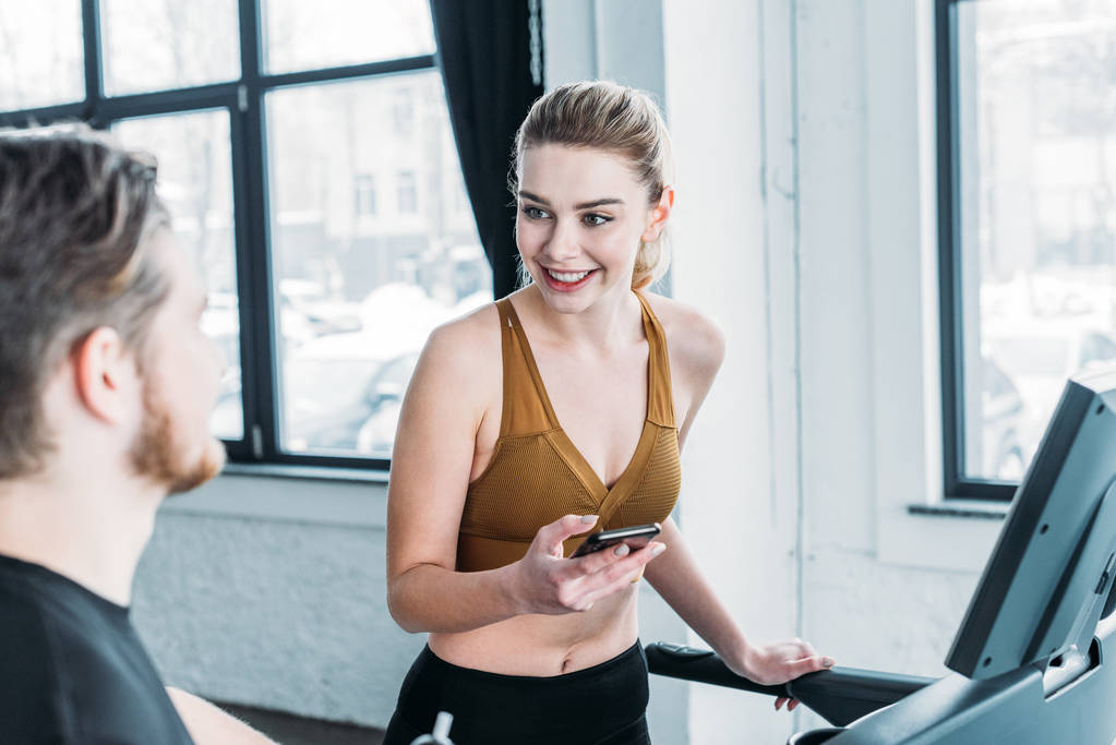 Uśmiechnięta dziewczyna ćwiczeń na bieżni z smartphone w rękę i patrząc na młody człowiek na pierwszym planie w siłowni - Zdjęcie, obraz