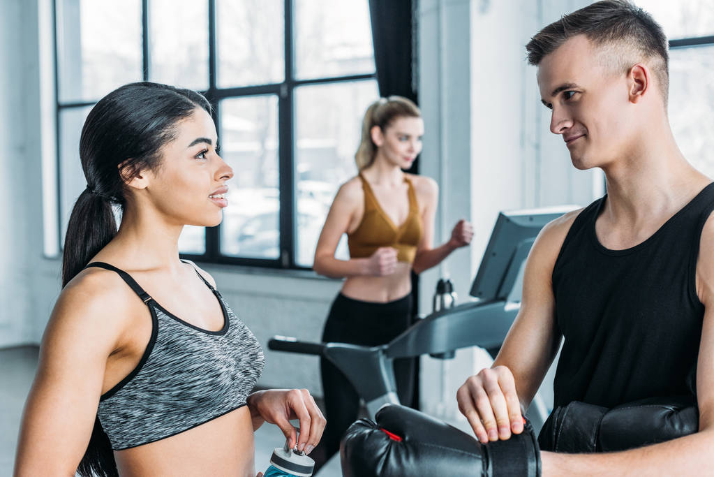 sportlicher junger Mann und Frau, die sich im Fitnessstudio unterhalten und anschauen, Sportlerin beim Training auf dem Laufband hinter sich - Foto, Bild