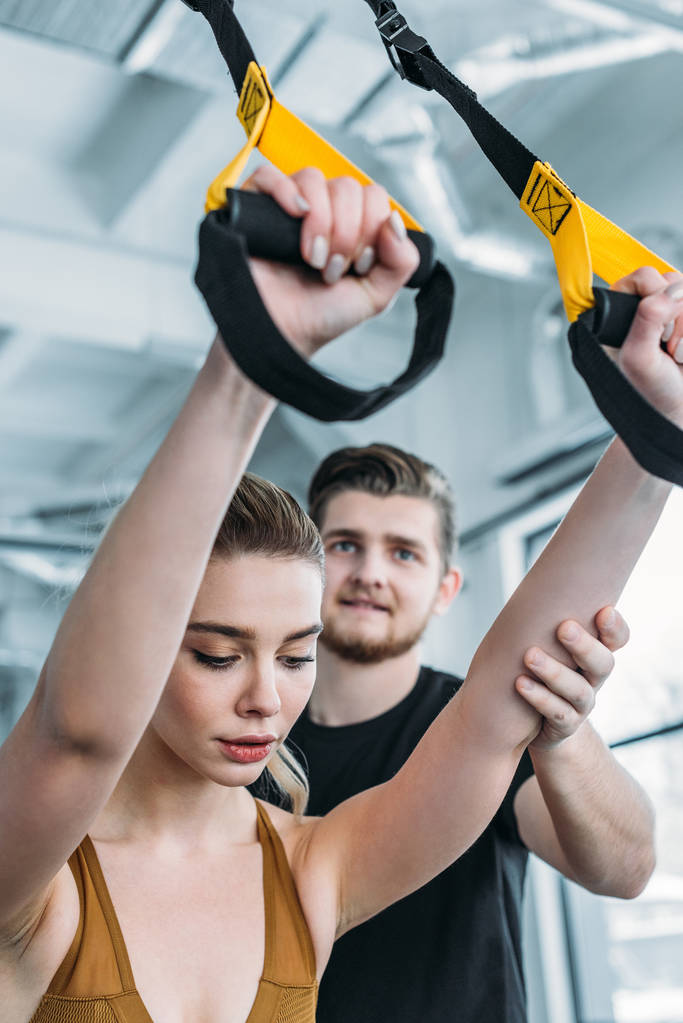 schöner lächelnder männlicher Trainer hilft attraktiven sportlichen Mädchen beim Training mit Aufhängebändern im Fitnessstudio - Foto, Bild