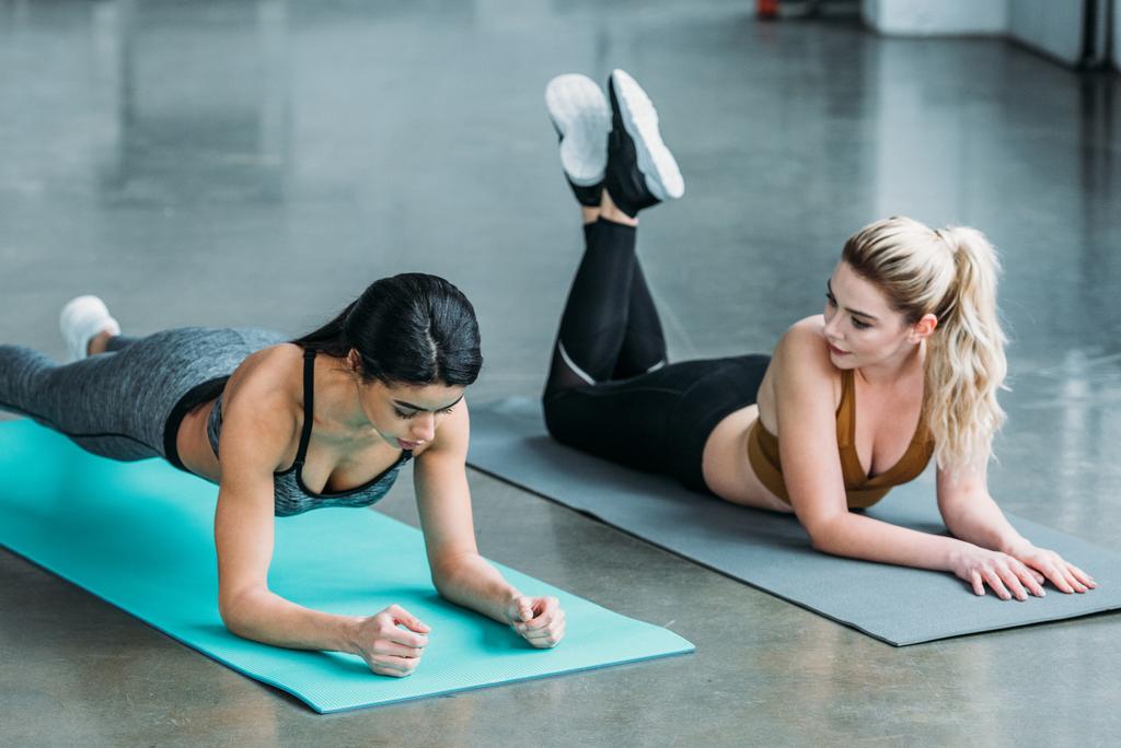 Blondes Mädchen liegt auf Yogamatte und sieht afrikanisch-amerikanische Sportlerin beim Plankentraining im Fitnessstudio an - Foto, Bild
