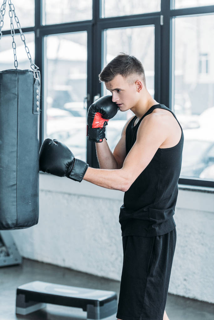 vista lateral del joven concentrado en guantes de boxeo haciendo ejercicio con saco de boxeo en el gimnasio
 - Foto, imagen
