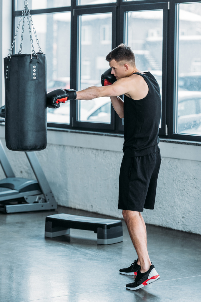 боковой вид молодого человека спортивного телосложения, боксирующего с сумкой в тренажерном зале
 - Фото, изображение