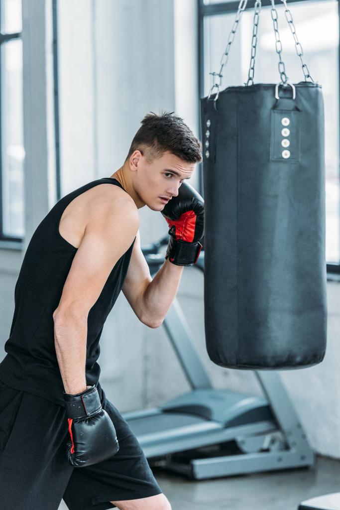 атлетичный юноша в боксёрских перчатках тренируется с боксёрской грушей в спортзале
 - Фото, изображение