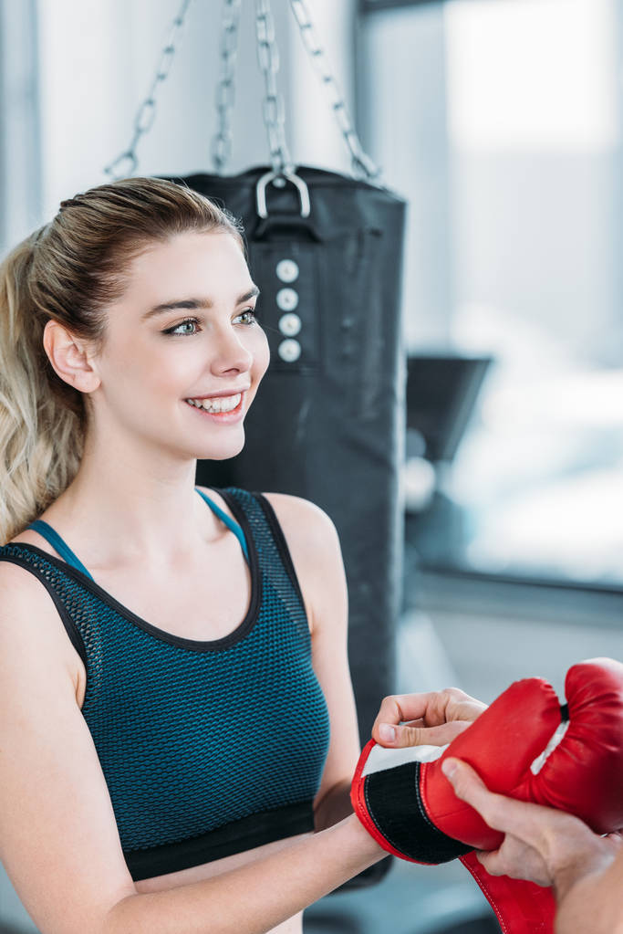 улыбающаяся юная спортсменка в боксёрских перчатках и смотрящая в спортзал
 - Фото, изображение