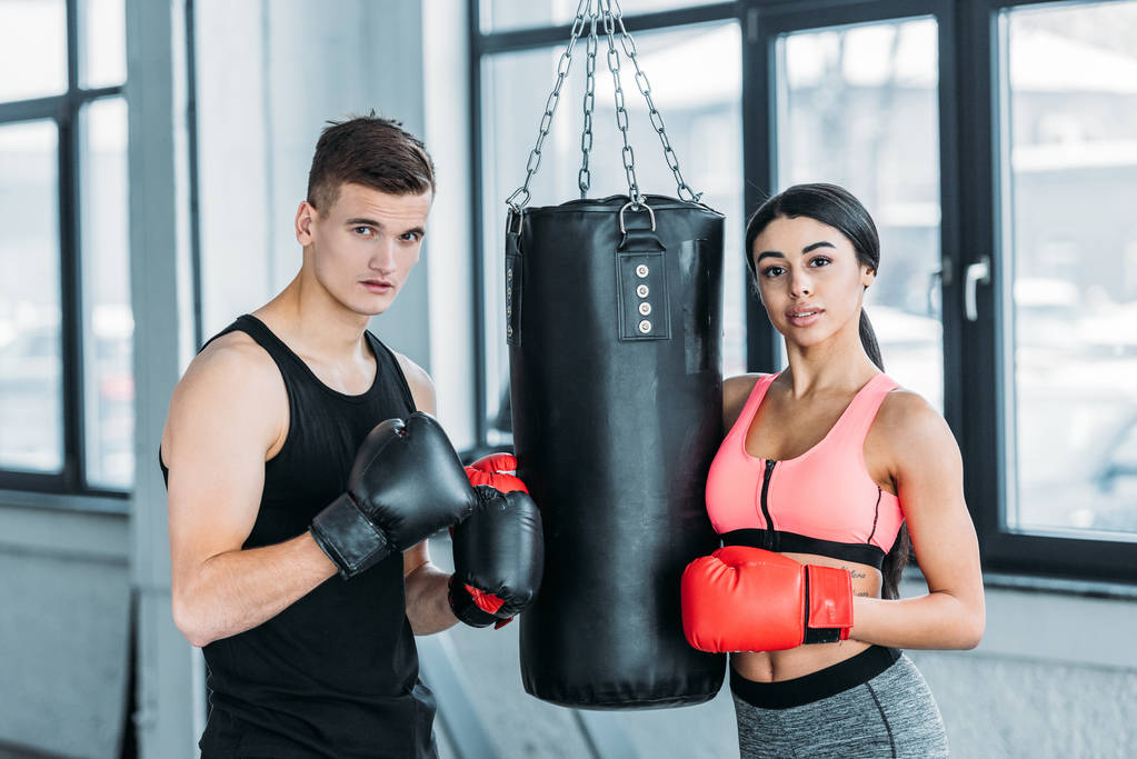 boxeadores masculinos y femeninos en guantes de boxeo de pie cerca del saco de boxeo y mirando la cámara en el gimnasio
 - Foto, Imagen