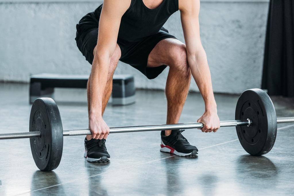 обрезанный кадр мускулистого молодого человека в спортивной одежде, поднимающего штангу в спортзале
 - Фото, изображение